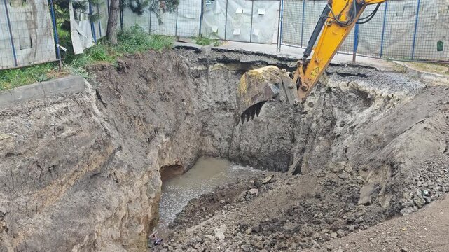 Yellow excavator bucket dumps and scrapes soil inside a deep excavation pit during public water supply pipeline repair to fix leaks in a pipe.