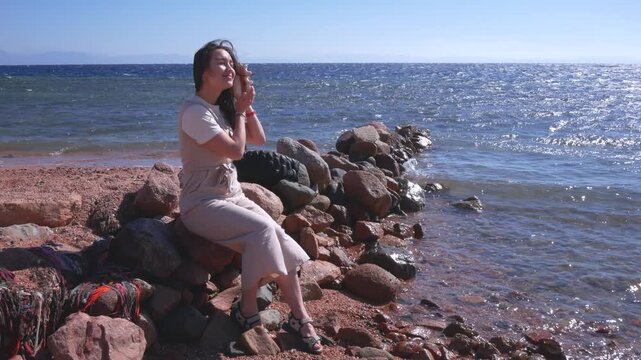 Young woman sitting on rocky beach shore listening to seashell ocean sound