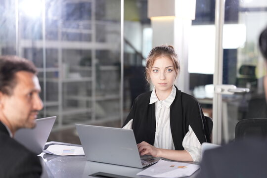 Businesswoman with a neat bun works at her laptop while a dark-haired male colleague sits opposite in profile during a focused meeting in a bright glass-walled coworking office.