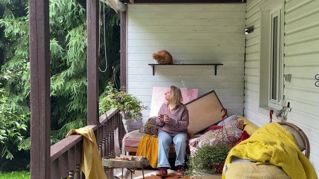 Woman sitting and drinking coffee on a cluttered veranda of a country house before makeover. Cozy outdoor space awaiting renovation with cushions, furniture, plants and home decor. Concept of porch 