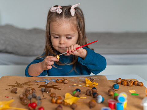 Cute toddler girl painting acorns with brush doing autumn DIY crafts