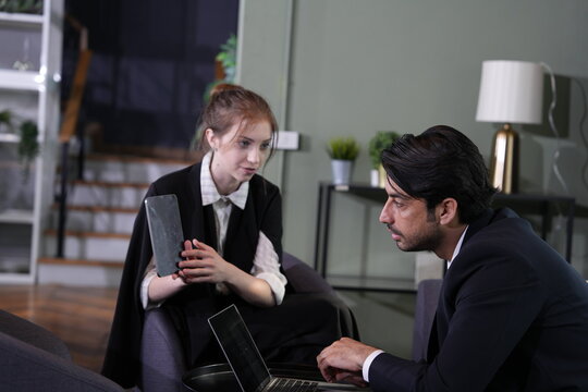 Bearded consultant frowns in concentration at the laptop while the young woman with the tablet sits back quietly in the opposite armchair, floor lamp glowing behind them.