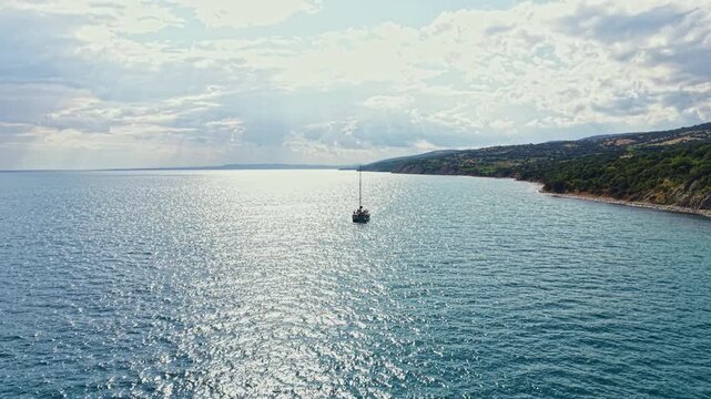 A sailing boat floats on clear blue water close to a rocky shore during a cloudy day. Waves gently lap against the hull as the coastline rises in the background, showing green trees and hills.