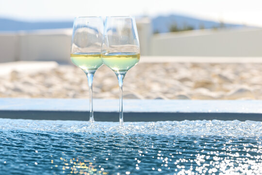 Two glasses of chilled white wine rest at the edge of an infinity pool in Santorini, reflecting sunlight and the serene elegance of a Mediterranean summer escape&mdash;perfect for a romantic island moment.