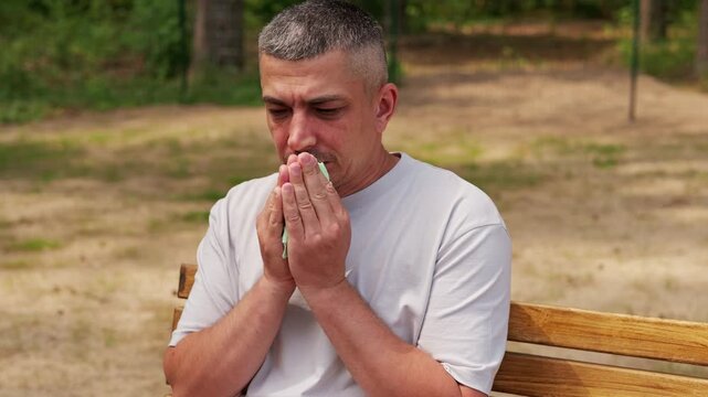 A man wipes his nose with a cloth while sitting on a wooden bench in a park. The sun is shining and the area is green with trees in the background