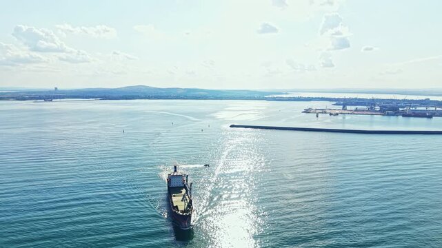 A cargo ship sails through water while leaving a wake behind. The sun reflects on the surface of the water. The harbor and land are visible in the background.
