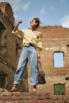 A stylish woman stands atop an old brick structure, wearing oversized jeans and a light shirt. The ruins are set against a clear blue sky, showcasing her confident posture and unique fashion choice