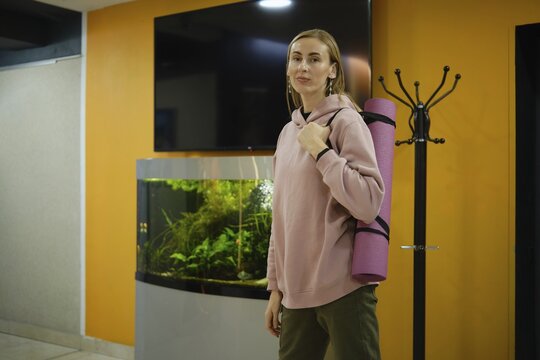 A woman in a pink hoodie stands in a fitness studio, holding a yoga mat. An aquarium with aquatic plants enhances the modern environment. The bright orange wall adds warmth to the space