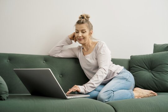 Middle-aged woman is comfortably seated on a green couch, focused on her laptop. She is wearing casual attire and appears engaged in her work, enjoying a relaxed moment in a cozy living area