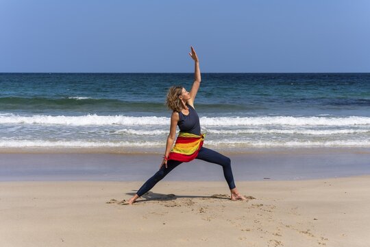 A woman doing yoga on a sandy beach by the sea. Shanti Virabhadrasana or Peaceful Warrior. Treen Cove, Cornwall, England, Great Britain