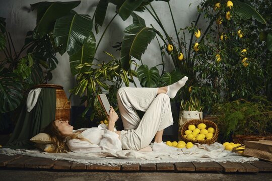 A woman is lying on the floor and reading a book. She is surrounded by plants and baskets of lemons. The setting is bright and full of greenery. The time appears to be during the day