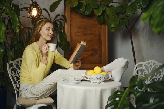 A woman sits at a table with her feet on the surface. She holds a cup and reads a book. A bowl of lemons sits on the table. Green plants surround her in a warm indoor setting