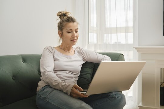 A woman sits comfortably on a green couch, focused on her laptop. She is learning new skills and concentrating on her task