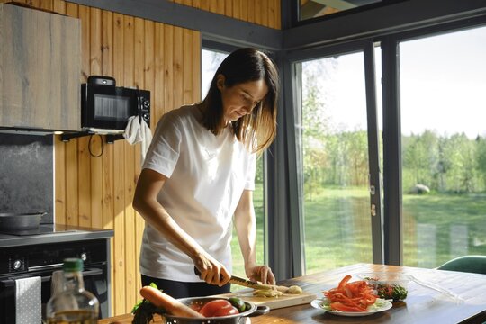 A woman is skillfully chopping ginger on a wooden table in a bright kitchen. Sunlight streams through large windows, highlighting fresh ingredients and a serene natural backdrop
