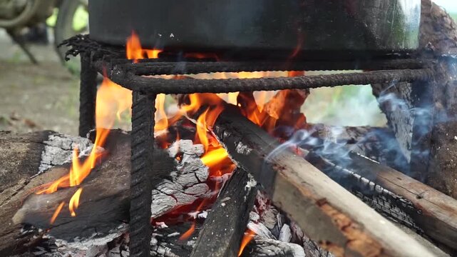 Traditional outdoor cooking over an open firewood campfire with bright orange flames under a large black metal pot on a rustic iron grill structure.