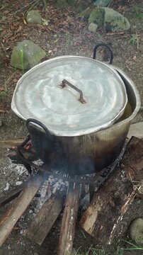 Large traditional aluminum pot with lid boiling over a rustic firewood campfire on an outdoor iron grill structure in the countryside.