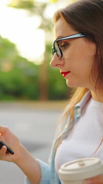 Happy woman with the smartphone and coffee to go stands on the street 