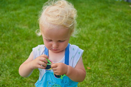 Little blond child playing with bright glasses on grass, Portrait of focused toddler twirling colorful glasses in hands