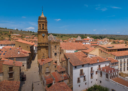 Aerial view of La Iglesuela del Cid showing the baroque church tower surrounded by medieval stone buildings, with a whitewashed arcaded building and narrow streets visible in the historic centre