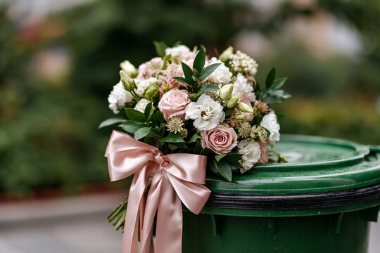 A small bridal bouquet with a satin bow in a trash bin outdoors. Wedding, ceremony, bouquet, romance, celebration, rustic, preparation, floristry, divorce, rejected love, broken heart
