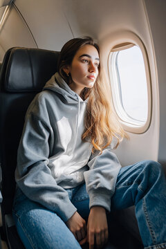 A young woman sits casually in an economy airplane seat, gazing out the window during a daytime flight. Travel, solo journey, contemplation, relaxation, lifestyle, aviation, modern travel, vacation