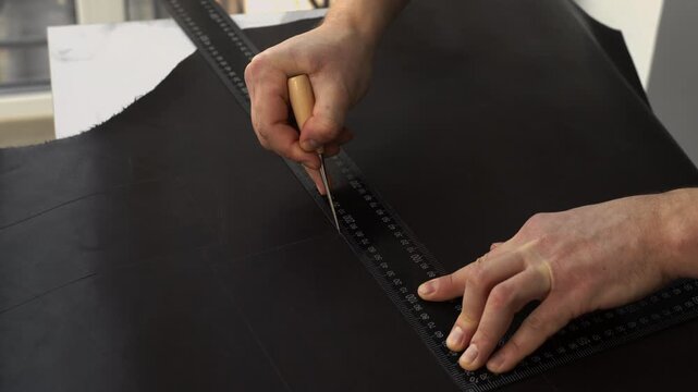 Artisan's hands meticulously cutting a piece of black leather using a traditional awl and ruler to create a precise, straight line