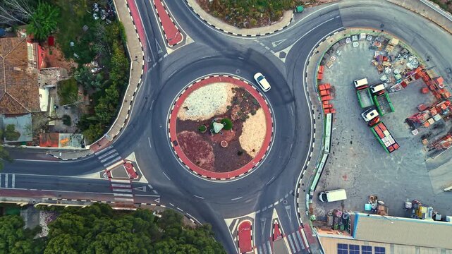 Vehicles move around the roundabout while construction work takes place nearby. Trees and bushes surround the traffic area as cars enter and exit from different roads.
