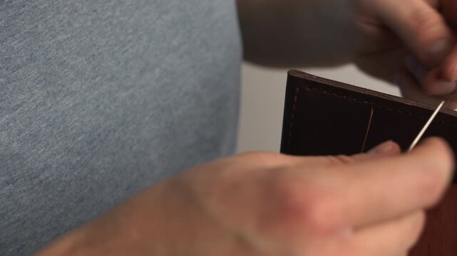 Artisan hand stitching brown leather wallet with needle and thread in a workshop creating handmade accessories