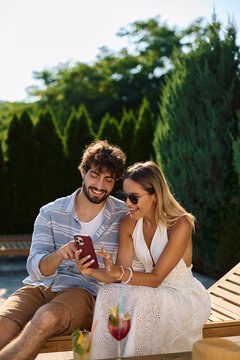 Happy couple using cell phone while relaxing by pool in summer day.
