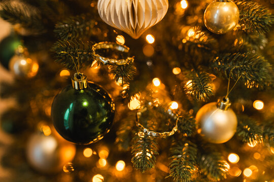 Close up of golden and green glass balls hanging on a Christmas tree with warm bokeh lights.