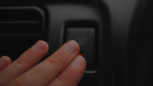 Fingers pressing the rear defroster button inside a vehicle, capturing automotive climate control, windshield visibility, winter driving, and road safety.