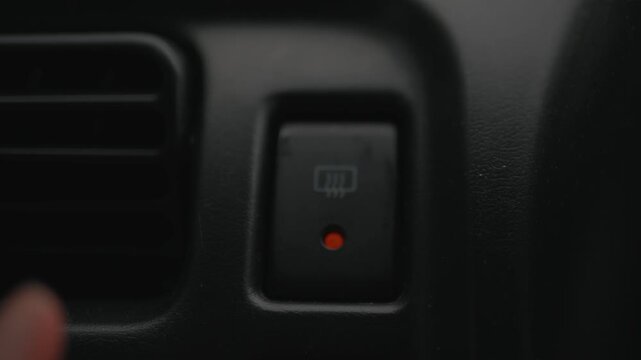 Close-up of a hand pressing the rear window defroster button on a car dashboard, representing vehicle climate control, visibility, driving safety, heating, and winter conditions.