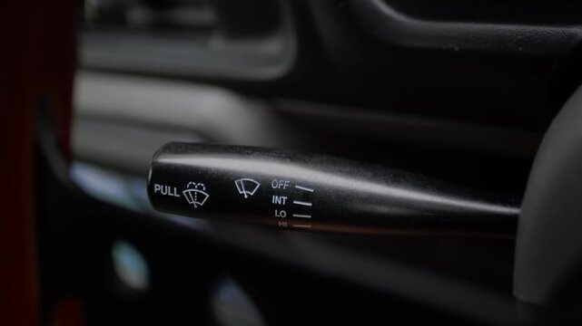 Close-up of a finger operating the windshield wiper control stalk in a car interior. Vehicle safety, driver control, maintenance, and rainy weather preparation.