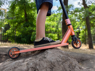 Fototapeta premium A boy stands with his feet on a stunt scooter stuck in a dirt hill in the park. The boy is preparing for a stunt.