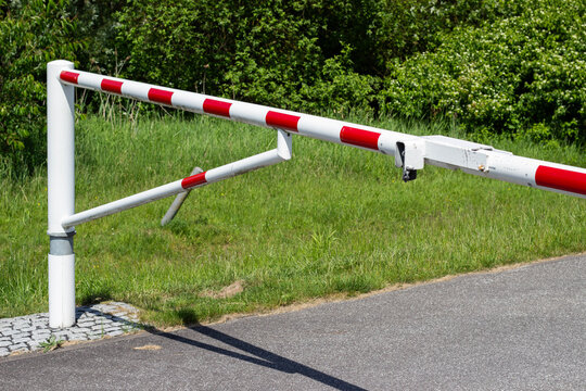 White and red barrier gate positioned at the entrance of a parking lot, surrounded by green grass and trees, with a paved road visible in the foreground