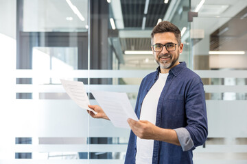 Fototapeta premium Confident businessman standing in a contemporary office corridor, holding various paperwork and smiling directly at the viewer, representing corporate success and professional communication