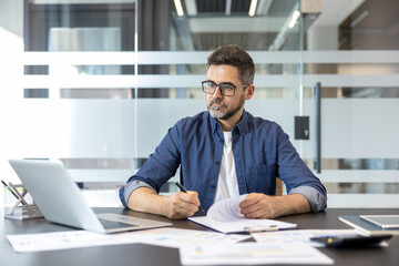 Obraz premium Man in a modern office concentrating on work, reviewing documents and writing notes while sitting at a desk with a laptop and various paperwork spread around