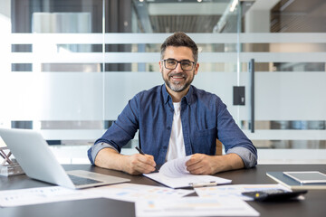 Obraz premium Confident businessman analyzing financial reports and signing paperwork, sitting at his desk with a laptop in a modern office, representing successful career and management