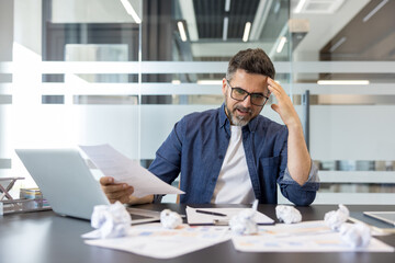 Obraz premium Businessman sitting at a desk in a modern office. Expressing frustration and anxiety while reviewing documents. Surrounded by a laptop and crumpled papers