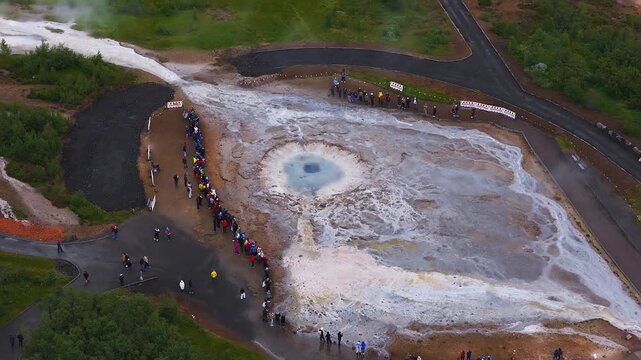 Aerial view of the Geysir geothermal area in Iceland shows the turquoise Geysir pool, steaming vents, mineral terraces, runoff channels, tourists, and walkways.