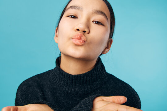Young woman with black sweater blowing kiss and looking at camera against blue background. Closeup portrait of female person sending love or affection with puckered lips and casual style.