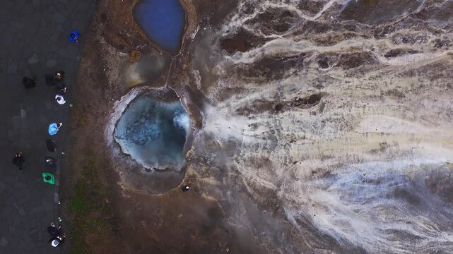 Aerial above Geysir geothermal area in Haukadalur, Iceland. Bubbling hot spring sends steam, people walk on paved path, sinter terraces and runoff spread right.