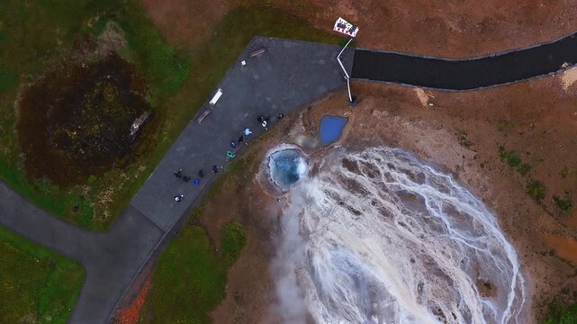 Aerial top view shows Geysir geothermal area, Haukadalur, Iceland. Tourists stand on a paved platform as steam rises and runoff flows from turquoise pools.