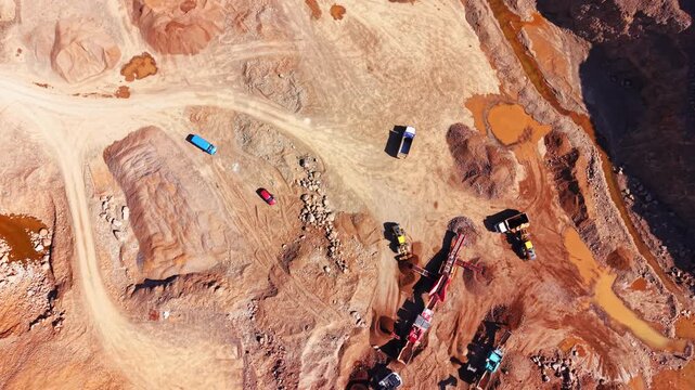 Quarry trucks and crusher machinery, Bulgaria. Overhead view of colorful vehicles on red dirt roads beside orange mineral ponds.