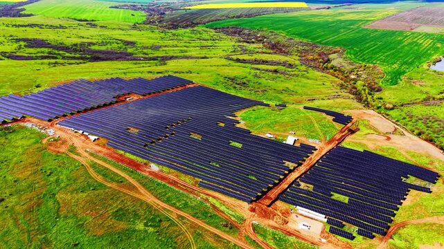 Aerial view of solar panel installation site. Dark photovoltaic modules bordered by green farmland and dirt path.
