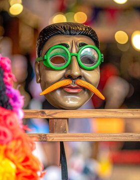 A traditional wax mask with green glasses and orange whiskers used in the Carnival of Binche sits on a shelf.