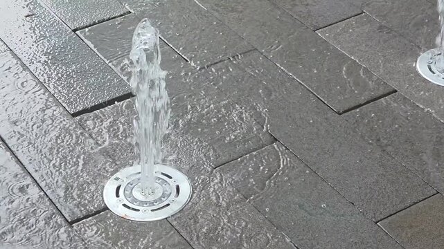 Water jets spray up from the ground in a public square during light rain in the city
