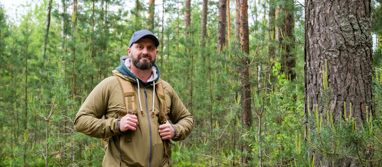 Man with backpack stands in pine forest and looks toward camera during hiking trip. Active recreation, exploration and travel lifestyle © Dmitrii Masquerade