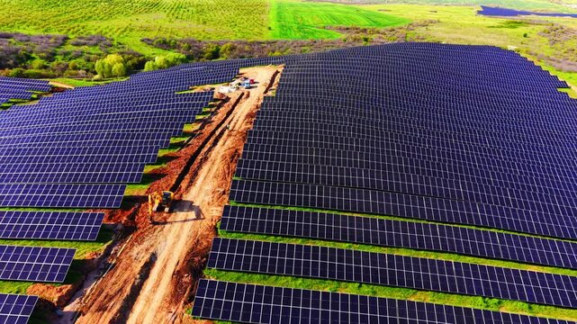 Construction of solar park. Unpaved dirt path running through rows of photovoltaic panels, heavy machinery parked in green fields during sunny development stage.