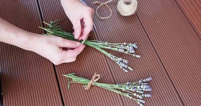 hands arranging lavender stems with string on wooden surface in focused and meticulous process top-down perspective, crafting home decor with brown and green tones.